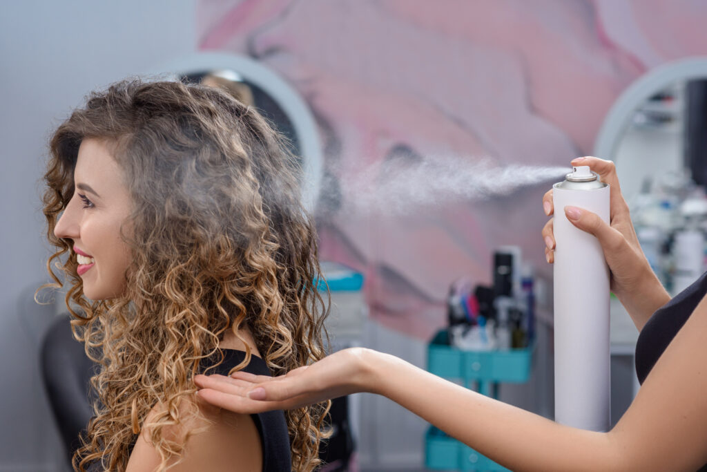 Stylist fixing hairstyle with hairspray in beauty salon