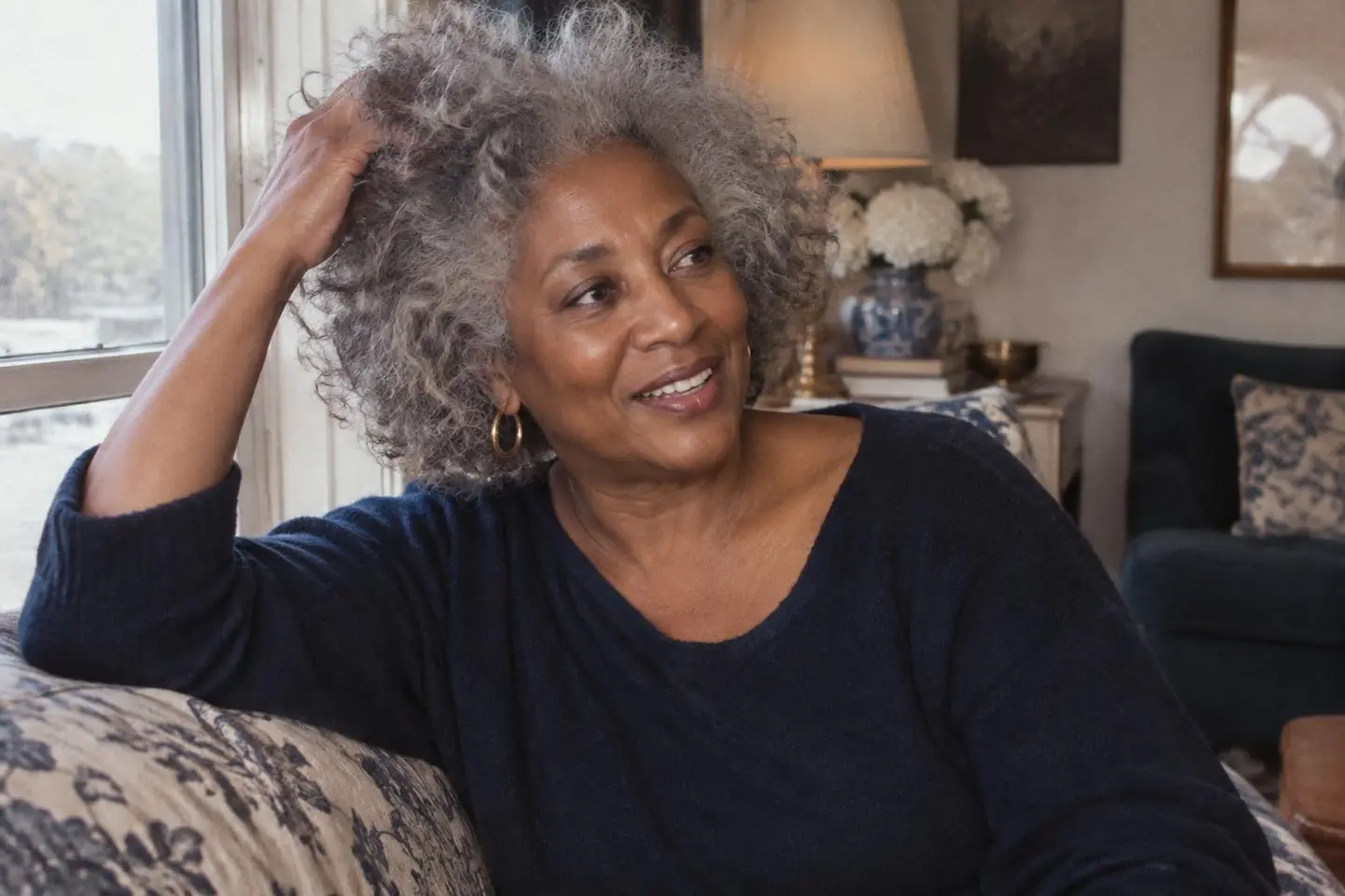 A Black woman lifting her naturally silver-gray curls by a window